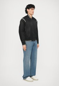 Fekete bombázódzseki cipzárral az elején, fehér csíkos díszítéssel, bordázott mandzsettákkal és szegéllyel. Széles szárú kék farmernadrággal és fehér sportcipővel viselve.