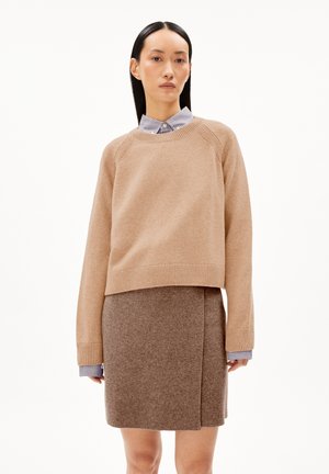 Pull en tricot beige avec un col rond, des bords côtelés et des épaules tombantes, superposé sur une jupe en laine marron avec une fente sur le côté.