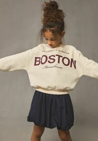 Krämfärgad oversized sweatshirt med "BOSTON" i vinrött, tillsammans med en marinblå skaterkjol. Har breda ärmar och ribbad nederkant. Subtil textur.