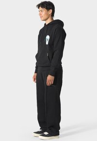 Hombre de pie de lado, vistiendo una sudadera negra con un pequeño gráfico en el pecho, pantalones de chándal negros con texto en el muslo y zapatillas negras con suelas blancas.