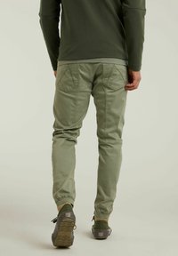 Person som går bortklädd i olivgröna joggarbyxor, matchande långärmad topp och mörkgröna sneakers med beige strumpor.