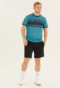 Sport T-shirt med korte ærmer i turkis med en marineblå stripe, parret med sorte shorts. Materialet ser let ud; enkel geometrisk mønster.