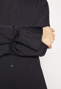 Sort skjorte med knaplukning, glat tekstur og plisserede ærmer. Har en klassisk krave og justerbar knaplukning på ærmerne.