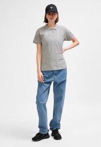 Grå pikétröja med krage, korta ärmar och en logga på bröstet, kombinerad med löst sittande blå jeans och svarta sneakers. Svart keps bärs.