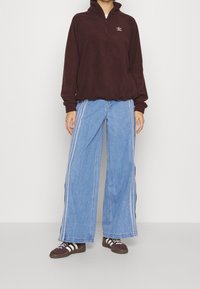 Bordeauxfärgad fleecetröja med dragkedja och hög krage, i kombination med vida ljusblå jeans med vita sidostycken och mörkbruna skor.