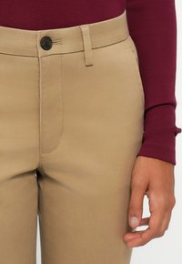 Pantalons beige ajustés avec une texture lisse, dotés d'une poche avant, de passants de ceinture et d'une fermeture à bouton foncé. Accompagnés d'un haut bordeaux à manches longues.
