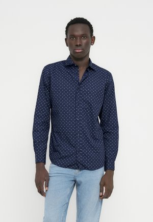 Jeune homme à la peau foncée portant une chemise à manches longues bleu marine à motif et un jean bleu clair, debout devant un fond blanc uni.
