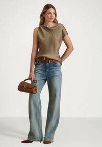 Moteris vilki aksominį rudą be rankovių viršų, plačius mėlynus džinsus, rudą diržą, rudus batus ir laiko mažą rudą rankinę.