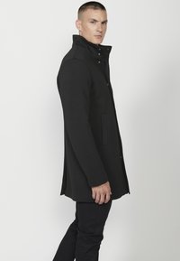 Cappotto in misto lana nero con colletto alto, tasche laterali e vestibilità sartoriale, caratterizzato da una texture liscia e un design minimalista.