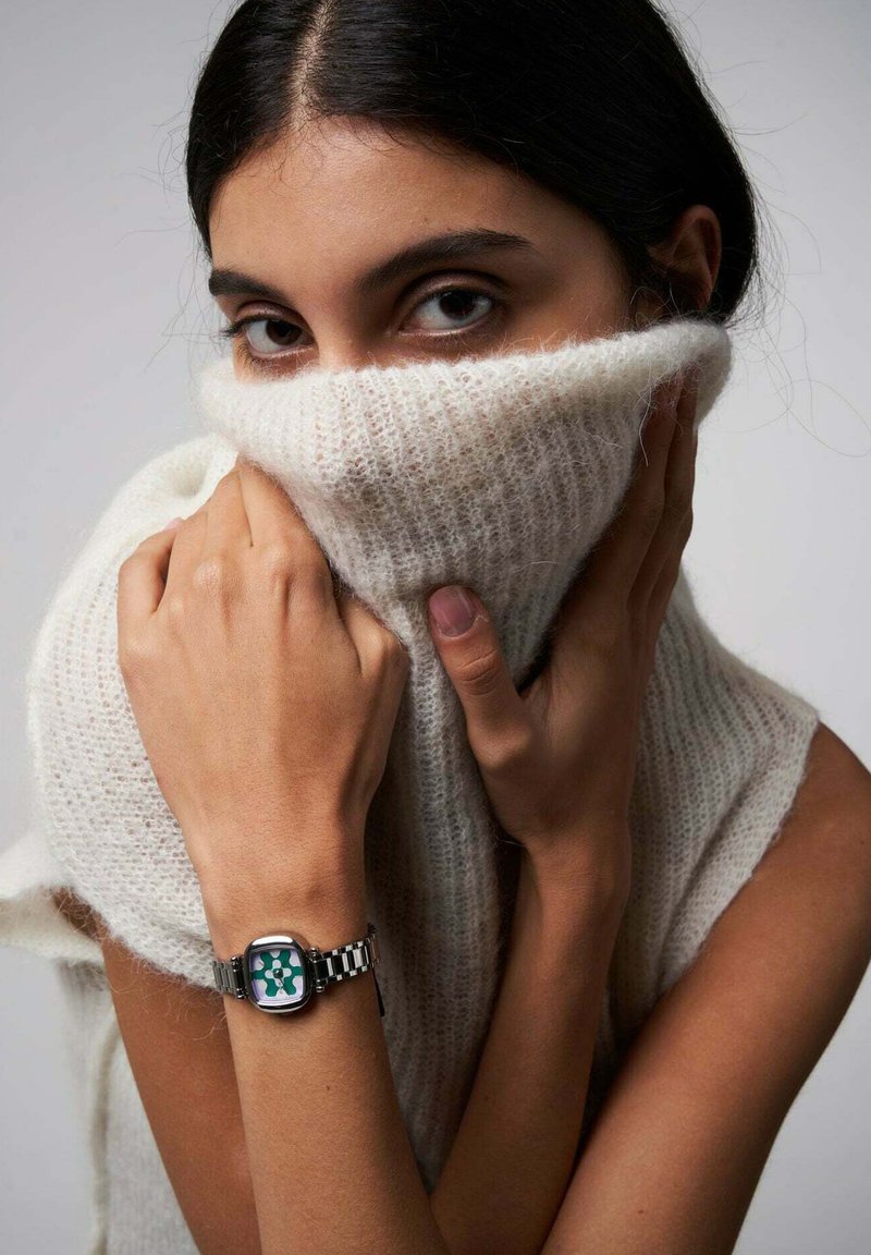 Montre-bracelet avec un cadran floral vert et blanc, boîtier en métal argenté, et un bracelet argenté et noir à motifs. Modèle portant un pull en laine crème.