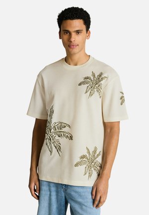 Man wearing a beige short-sleeve shirt with green palm tree prints, paired with light blue jeans, standing against a white background.