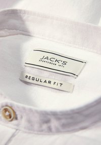 Interno di una camicia bianca con colletto, caratterizzato da un'etichetta in tessuto con testo nero "JACK'S SPORTSWEAR INTL" e "REGULAR FIT", accentuato da un bottone.