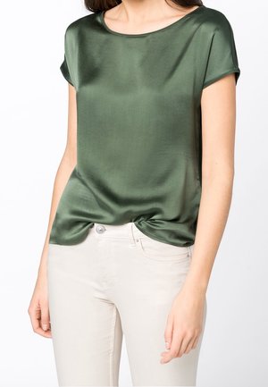 Groene satijnen top met korte mouwen en een ronde halslijn, een gladde textuur en een knoopdetail aan de onderkant, gecombineerd met lichtgekleurde jeans.