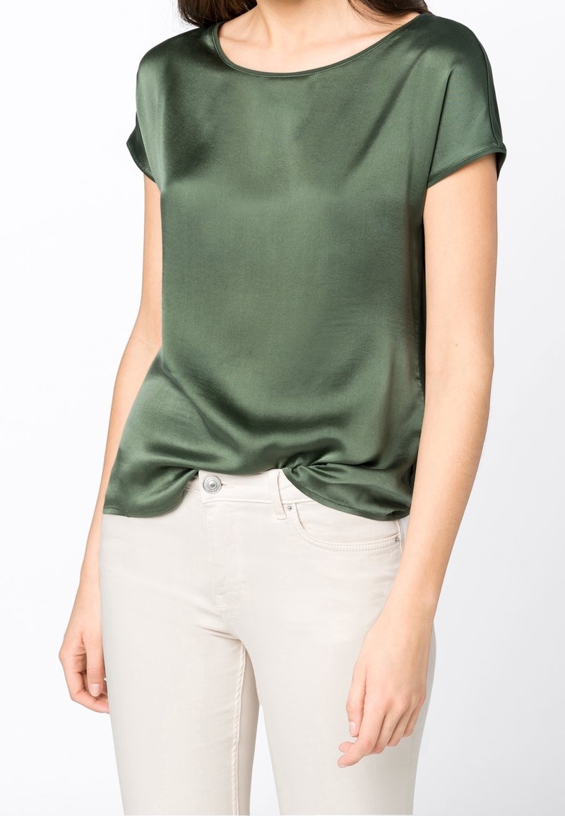 Haut court en satin vert à manches courtes avec un col rond, texture lisse et un détail noué à l'ourlet, associé à un jean de couleur claire.