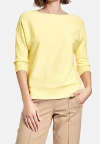 Gul stickad tröja med vid halsringning och tre fjärdedelars ärmar. Har en ribbad nederkant och ärmslut, i kombination med beiga byxor.