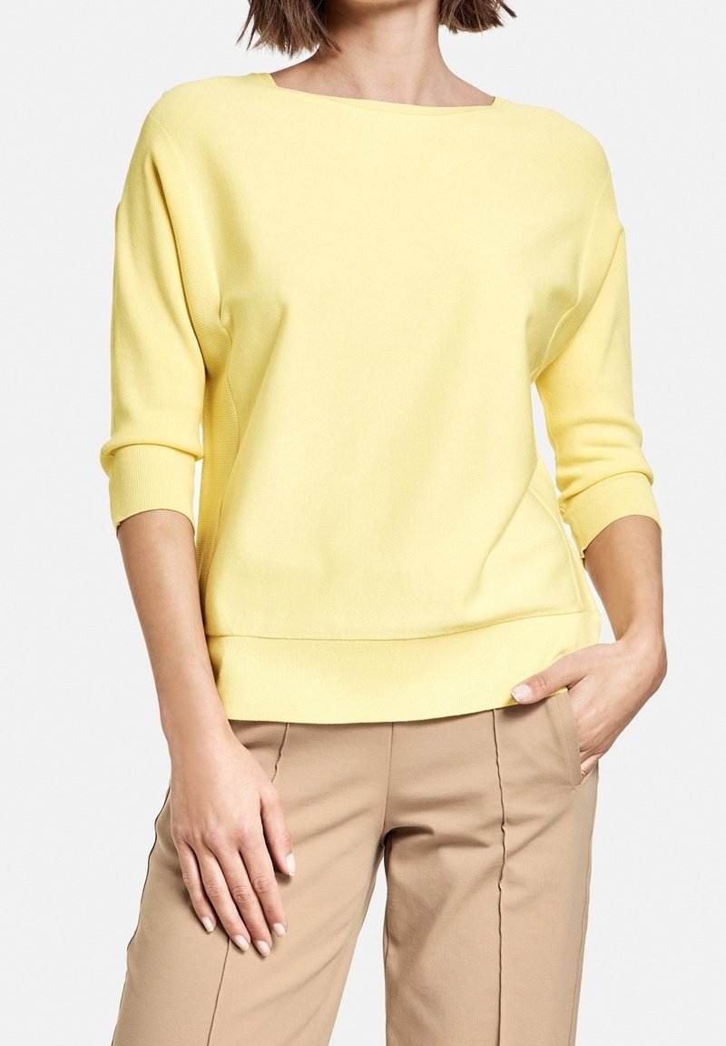 Gul stickad tröja med vid halsringning och tre fjärdedelars ärmar. Har en ribbad nederkant och ärmslut, i kombination med beiga byxor.