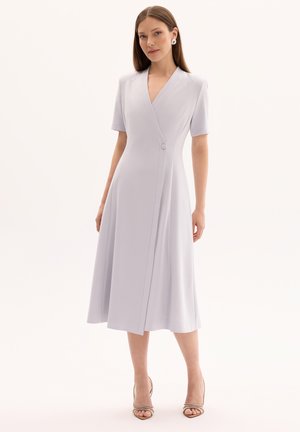 Kobieta w jasno lawendowej, midi sukience z krótkim rękawem, o kopertowym kroju i pojedynczym guzikiem, w połączeniu z beżowymi sandałami na wysokim obcasie.