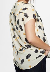 Blouse à manches courtes en tissu crème clair ornée d'un motif ananas noir et d'étoiles orange. Coupe décontractée avec un ourlet arrondi.