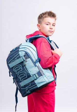 Chlapec v červené mikině s kapucí nese velký modro-zelený batoh přes jedno rameno a dívá se do kamery na jednobarevném pozadí.