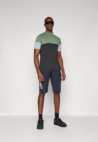 Camisa polo de hombre en verde menta y carbón con mangas cortas, combinada con pantalones cortos azul marino con un logo blanco, llevada con zapatos negros y gafas de sol.