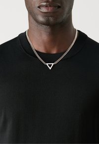Collier en chaîne argentée avec un pendentif triangulaire au centre noir, porté sur un t-shirt noir à col rond. Texture lisse, design moderne.