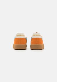 Oranžové semišové tenisky s modrými akcenty a béžovým patním jazykem s zlatým logem "Reebok". Viditelná texturovaná gumová podrážka.