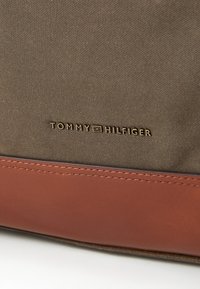 Brown fabric bag with a smooth leather bottom. Features gold embossed "TOMMY HILFIGER" logo. Textured surface with visible stitching detail.