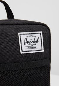 Sac en tissu noir texturé avec une étiquette rectangulaire blanche de Herschel Supply Co. cousue sur le côté supérieur avant.