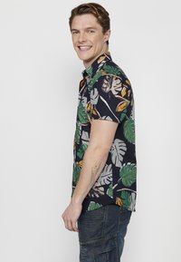 Camisa de manga corta con botones que presenta un fondo navy con grandes patrones de hojas verdes y amarillas, confeccionada en una tela ligera.