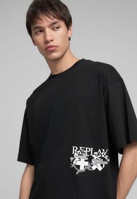 T-shirt in cotone nero con vestibilità comoda, scollo rotondo e stampa grafica sul lato sinistro con la parola "REPLAY" e design astratti.