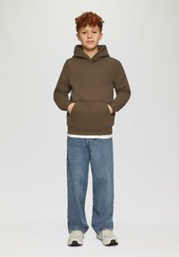 Felpa marrone con tasca frontale e testo "CONFIDENT" sul cappuccio, abbinata a jeans a gamba larga blu chiaro e sneakers grigie.