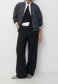 Grijze rits sweatvest, zwarte tanktop, witte blouse, brede zwarte broek, platte schoenen met zilveren accenten. Soepele stoffen en een relaxed fit.