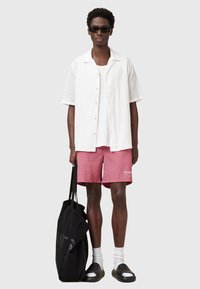 Hvid, tekstureret kortærmet skjorte, ensfarvet hvid tanktop, lyserøde shorts med logo, sort tote bag, sorte solbriller, hvide sokker, sorte slidesandaler.