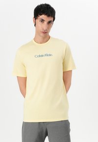 Camiseta de algodón amarilla con mangas cortas y cuello redondo, que presenta un logotipo gráfico azul "Calvin Klein" en el pecho.