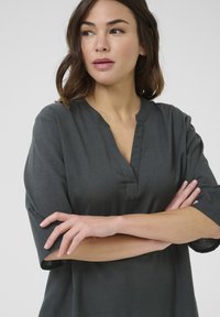 Tmavě šedá tunika s krátkými rukávy, výstřihem do V a jemnou texturou. Má volný střih a ohrnuté manžety rukávů.