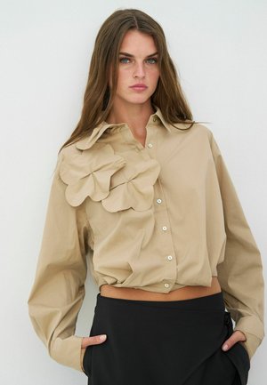 Beige skjorte med knapper og store stofblomsterdetaljer på venstre side, med cropped design og lange ærmer.