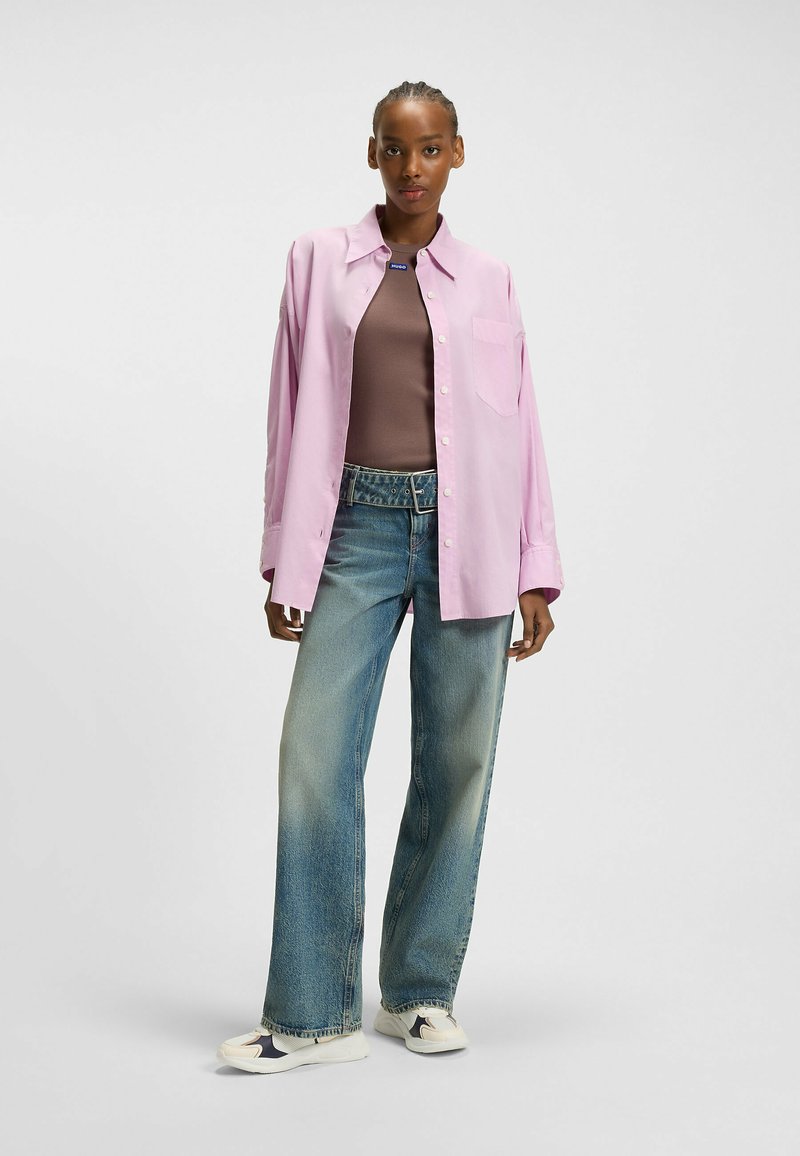 Helles rosa, übergroßes Hemd mit Knöpfen, das über einem braunen, figurbetonten Oberteil getragen wird, kombiniert mit weit geschnittenen, ausgewaschenen blauen Jeans und Sneakers mit Akzenten.