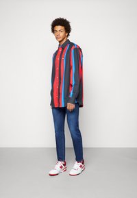 Pasiasty, button-up koszula w kolorach czerwonym, niebieskim i szarym; luźny krój; w połączeniu z niebieskimi dżinsami i białymi trampkami z czerwonymi akcentami.