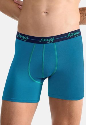 Mann trägt türkisfarbene Boxershorts mit dunkelblauem Bund, der mit "Sloggi" gebrandmarkt ist, zeigt den Mittelteil und die oberen Oberschenkel vor weißem Hintergrund.