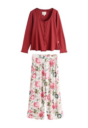 Frau in rotem Oberteil und geblümter Hose sitzt barfuß auf einem weißen Metallstuhl neben einem weißen Tisch mit Teekanne und Blumen auf einer Backstein-Terrasse.
