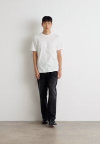 Weißes Baumwoll-T-Shirt mit einem dezenten Druck, kombiniert mit einer schwarzen Jeanshose und schwarzen Schuhen, stehend vor einer einfarbigen Wand.