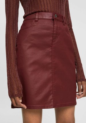 Gros plan d'une personne portant une jupe en cuir bordeaux brillante avec des passants de ceinture et un bouton à l'avant, associée à un haut en maille scintillante à manches longues.