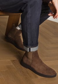 Stivali Chelsea in camoscio marrone con finitura liscia, pannelli laterali elasticizzati e suola nera a profilo ribassato, abbinati a jeans scuri in denim arrotolati.