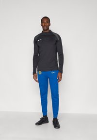 Czarna, długosleeve'owa koszulka sportowa z kołnierzykiem i szarymi akcentami, zestawiona z niebieskimi spodniami z logo oraz czarnymi butami sportowymi.