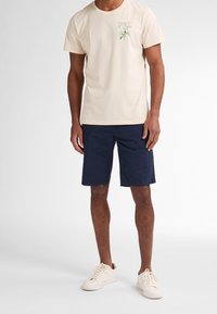 Camisa de algodón beige con un pequeño gráfico verde y rosa en el pecho, combinada con pantalones cortos azul marino. Zapatillas blancas completan el atuendo.