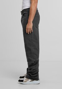 Muž v bledých černých džínách a bílých, černých a béžových teniskách stojící bokem proti jednoduchému pozadí.