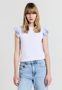 Top bianco a coste con maniche a volant in pizzo blu, abbinato a jeans azzurri a vita alta. Collo rotondo semplice, vestibilità comoda.