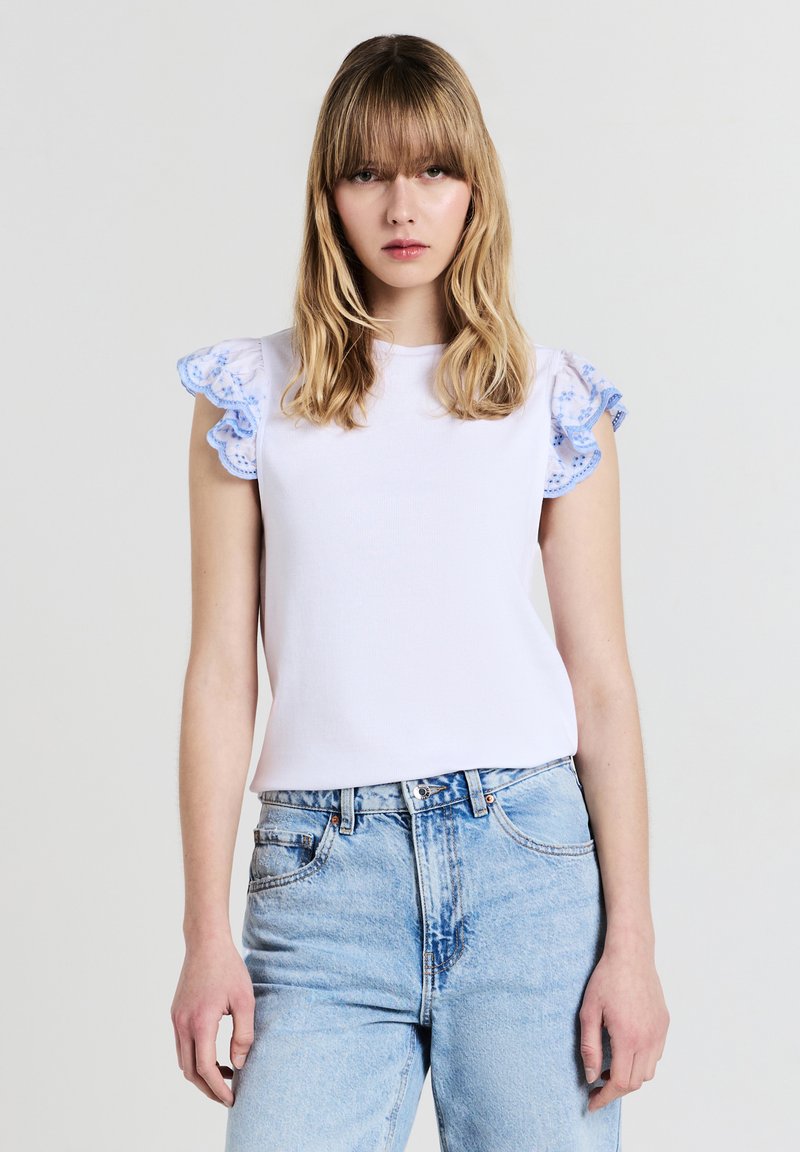 Top bianco a coste con maniche a volant in pizzo blu, abbinato a jeans azzurri a vita alta. Collo rotondo semplice, vestibilità comoda.