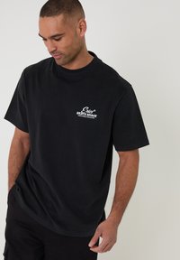 Hombre con camiseta negra de "Etnies Skate Shack Santa Monica Boulevard", mirando hacia abajo, con las manos parcialmente en los bolsillos, frente a un fondo gris claro.