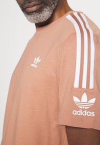 Kortärmad korallfärgad t-shirt i bomull med en vit Adidas-logotyp på bröstet och tre vita ränder på axlarna.